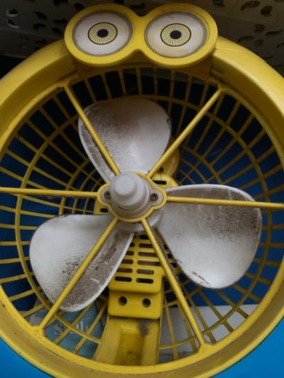 This is a close-up, eye-level, medium shot of a yellow, plastic fan with a cartoon-like design. The fan has a prominent yellow casing and a blue grate behind the fan blades. The fan blades are white and appear dusty. On the top of the fan casing, there are two decorative "eyes" with black and yellow irises, resembling a minion character. The fan is angled slightly upwards. The background is dark and out of focus, showing some textured surface. This image was taken in Karangkemiri, Indonesia.