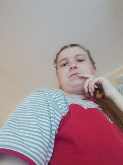 The image shows a low-angle, close-up shot of a young person wearing a red shirt with grey and white striped sleeves. The person has a pigtail and is resting their chin on their hand, with their finger touching their lips. Their eyes are looking towards the camera, and they have a thoughtful or contemplative expression. The background is a plain, light-colored wall. The lighting appears to be indoor, with no specific indicators of time of day or weather. There is no visible text in the image. The overall impression is of a casual, personal photograph.