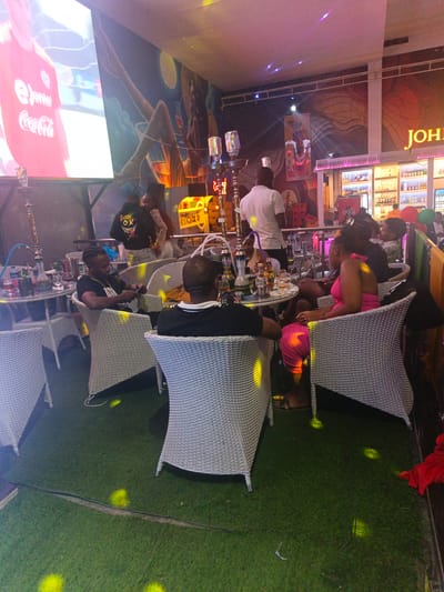 **Location:** Syokimau-Mulolongo ward, Kenya.
**Scene:** An indoor establishment with multiple groups of people seated around tables.
**People:** Several individuals are present. One person is standing wearing a white shirt. A person in a pink outfit is seated at a table. Other individuals are either seated or standing around tables, engaged in conversation and activities.
**Subjects:** The primary subjects are the people within the establishment.
**Objects:** Prominent objects include hookahs, tables, chairs, alcoholic beverages, a television screen displaying a Coca-Cola advertisement. There is a bar in the background.
**Actions:** People are socializing, consuming drinks, and watching a television screen. One individual is standing near a table.
**Interactions:** People are interacting with one another at their respective tables.
**Environment:** The environment is a dimly lit indoor establishment. The floor appears to be covered in artificial grass.