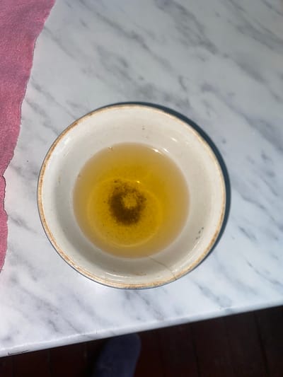 A close-up, overhead shot depicts a small, shallow, off-white ceramic bowl filled with a light golden-colored liquid, likely tea. The liquid has a slightly cloudy appearance, with a dark, circular concentration of sediment at the center. The bowl itself is rimmed with a narrow band of what appears to be dark blue, with a thin, worn brown line beneath it. The bowl rests on a white marble countertop with gray veining. To the left of the bowl, a portion of a pink, textured fabric, possibly a towel, is visible. The lighting suggests an indoor setting. The image was taken in Dashtabad, Uzbekistan.