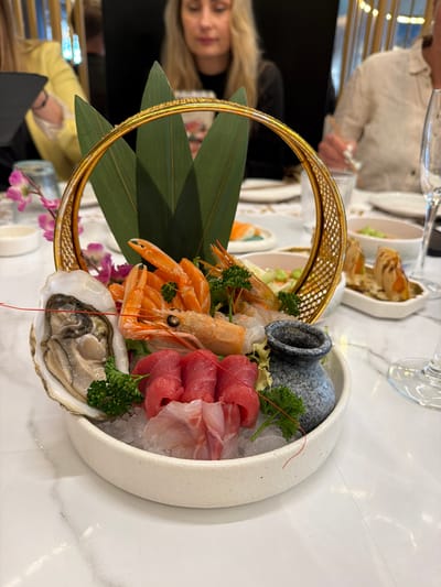 The image depicts a decorative platter of seafood, arranged artfully in a white bowl. The platter contains oysters, shrimp, and sliced raw fish, likely sashimi. These items rest on crushed ice and are garnished with greenery and a small grey decorative pot. A golden circular frame surrounds part of the platter, with large green leaves adding to the presentation.

The setting appears to be a restaurant. Parts of other diners and tables are visible in the background, including someone wearing a light-colored sweater and another in yellow. Silverware and additional dishes suggest a meal in progress.

There is no information available within the provided image to confirm the location as Torreforta, Spain.
