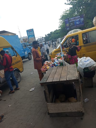 The image depicts a busy street market scene in Satellite Town, Nigeria, during daytime under an overcast sky. The setting is an urban environment with vehicular and pedestrian traffic.
In the foreground, a wooden market stall, constructed from crates, holds various packaged snacks, bottled drinks, eggs, and bread. A woman, likely a vendor, wearing a colorful patterned dress and headscarf, stands behind the stall, appearing to arrange or attend to the goods. To her left, a man in an orange traditional outfit observes the stall, with another man in a striped shirt and black cap standing nearby.
Several vehicles are present. A prominent yellow mini-van, commonly used for public transport, is parked near the vendor's stall, with people visible inside. Further to the left, another yellow van is being loaded or unloaded by a man in a red patterned shirt and blue jeans. A blue truck and other vehicles are seen further down the street, indicating a bustling thoroughfare. People are also walking in the background.
Notable details include the paved ground, scattered litter, and large billboards rising above the street. One billboard advertises "The best product." Another prominent billboard for "JP BATTERIES" states "NOW IN ASPANDA," offering "MORE POWER, LONGER LIFE, GUARANTEED PERFORMANCE," with "12 MONTHS WARRANTY" and "15 MONTHS WARRANTY" seals, a phone number "0814 877 7630," and website "WWW.JPCHRIS.COM," along with an address mentioning "ASPANDA TRADE FAIR." A white plastic bag near the stall has the partially visible text "Lotrec." The overall atmosphere is one of active commerce and daily urban life.