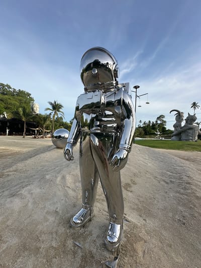 A large, chrome-plated sculpture of an astronaut stands in the foreground on a sandy area. The astronaut sculpture is highly reflective, showing distorted reflections of the surrounding environment, including trees, sky, and other sculptures. In the background, there are palm trees, a large sculpture of a face on a pedestal, a large silver sphere, and other abstract sculptures. The scene appears to be an outdoor art installation or sculpture park. The location is identified as Baan Lipa Noi, Thailand. The sky is blue with some scattered clouds. The lighting suggests it is daytime, likely late afternoon or early morning due to the low angle of the sun's light and the long shadows. There are no people visible interacting with the astronaut sculpture in the foreground.