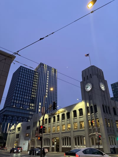 This image captures a street-level view of the San Francisco Chronicle building in San Francisco, California, USA, at dusk. The iconic building, characterized by its distinctive art deco architecture, features a tall clock tower with illuminated clock faces. An American flag flies from the top of the tower. To the left of the Chronicle building, two modern high-rise buildings rise, their windows reflecting the twilight sky. The street in front of the buildings is wet, indicating recent rain, and several cars are visible, some with their headlights on. Traffic lights are illuminated, showing red and yellow signals. Overhead, power lines crisscross the sky, and a streetlamp casts a warm glow. The sky is a deep twilight blue, with a faint hint of a moon. The overall impression is one of an urban environment transitioning from day to night.