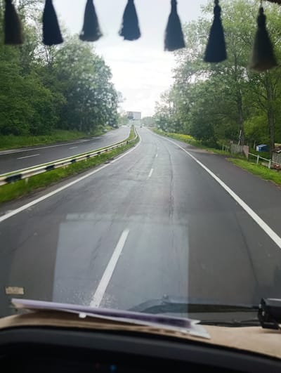 The media file depicts a view from inside a vehicle traveling on a wet asphalt road. The road curves to the left, with two lanes in each direction separated by white lines. On the left side of the road, a metal guardrail runs alongside a grassy embankment. Dense green trees and foliage line the road on both sides. In the distance, a billboard is visible on the right side of the road, featuring a portrait of a man. Further down the road, several vehicles are seen. The sky is overcast and appears to be a hazy, bright gray. The windows of the vehicle have water droplets on them, suggesting it might be raining or has recently rained. The foreground shows the dashboard of the vehicle, with papers resting on it, and a glimpse of the steering wheel. The overall scene is that of a journey on a rural or suburban road on a cloudy day. There is no discernible text in the visible area apart from the potential text on the billboard.