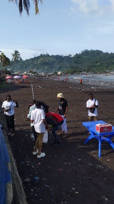 The scene is a beach in Limbe, Cameroon, located at coordinates 4.004 latitude and 9.211 longitude. There are seven people on the beach. Four are standing and three are seated. The people are all wearing casual clothing. One person is wearing a white shirt and black pants, another is wearing a white shirt and khaki pants, and another is wearing a white shirt and blue jeans. One person is wearing a red shirt. There are several palm trees on the beach. In the background, there are buildings and a forested hillside. The ground is covered in dark sand. There is debris and litter scattered across the beach. A blue table with a red box on top is on the beach.