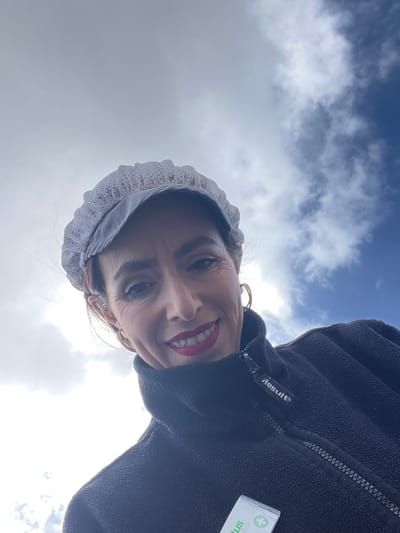 A woman wearing a white bonnet-style head covering and a black fleece jacket with the brand "Result" visible, is looking down at the camera. She has fair skin, dark eyebrows, and red lipstick. A small rectangular object with a green cross is visible on the jacket. The background shows a partly cloudy sky with blue patches. The image appears to be taken outdoors, possibly during the day, but the location in Roodt-sur-Syre, Luxembourg, cannot be determined from this image.
