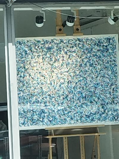 A large, predominantly blue and white artwork covers a frame. The artwork appears to be made of small, fragmented pieces. Three spotlights are positioned above the artwork, illuminating it. The artwork rests on a wooden easel. The surrounding area includes reflective surfaces and structural supports.