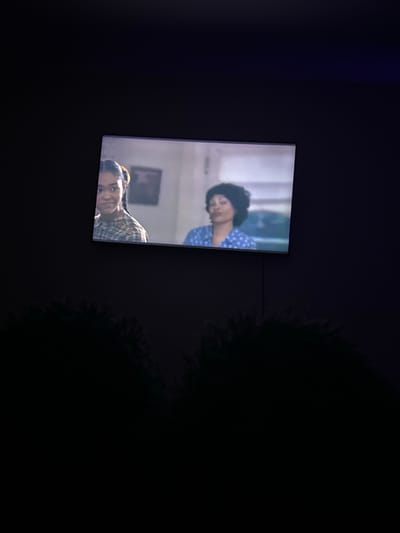 The image displays a television screen showing a scene with two individuals. The top half of the screen features a person with dark, curly hair wearing a blue and white polka dot shirt, their head tilted downwards. Below them, another person is visible, looking towards the camera with their head resting on their hand. This second individual is wearing a plaid shirt. The background on the screen appears to be an indoor setting with hints of furniture and walls. The overall image is dimly lit, suggesting it might be nighttime or a dark room. The television screen itself is the primary illuminated object, casting a faint glow. The context of Sof'ino, Russia is provided, but no specific visual cues within the image directly indicate this location or any particular weather conditions. No text is visible in the media file.