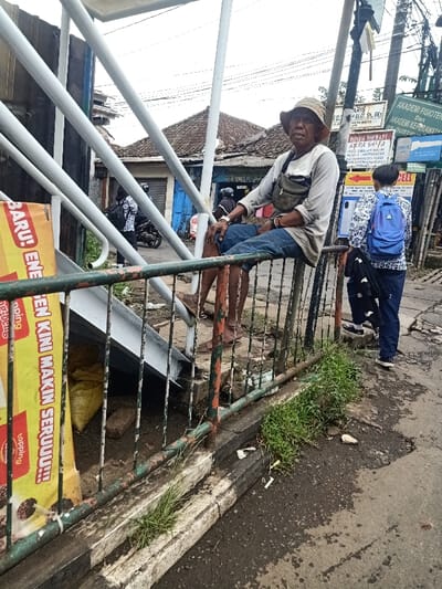An elderly man wearing a hat and casual clothes sits on a rusted green railing next to a pedestrian overpass. Adjacent to him, a person with a blue backpack stands on the sidewalk. Behind them are signboards with various inscriptions, including "AKADEMI FISIOTERAPI" and "RUMAH THERAPI". A motorbike is parked in the background, and there's a banner attached to the overpass railing with the text "BARU! ENE BEN KINI MAKIN SERUUU!!!". The street appears damp, and there is vegetation growing along the edge of the sidewalk.