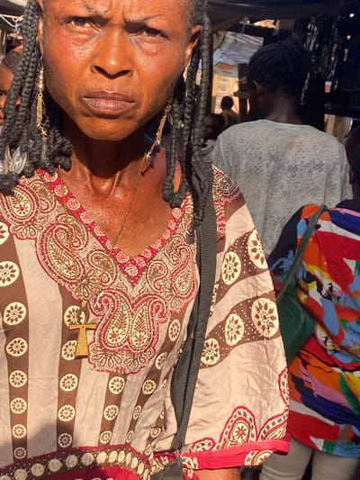 The image depicts a close-up, waist-up view of an adult Black woman with a stern expression, captured in Amaseri, Nigeria. She has a dark complexion, pronounced wrinkles on her forehead and under her eyes, and long, dark hair styled in numerous individual braids or dreadlocks. Her eyes are dark and gaze slightly to the viewer's left. Her lips are full and slightly parted. She wears a gold-colored chain necklace with a prominent golden Ankh pendant resting on her chest, and two long, dangling gold-colored earrings with chain links and star-shaped charms. Her attire is a V-neck tunic or dress featuring a cream and red paisley pattern around the collar and chest. The main body of the garment has vertical alternating stripes of light beige and brown, with the brown stripes containing circular, flower-like motifs. A black strap, likely from a bag, is visible over her left shoulder.
In the mid-ground, behind the main subject, another individual is seen from the back, wearing a light-colored, patterned long-sleeved top, possibly white or cream lace. Their dark hair is pulled back. To the far right, a third person is partially visible, wearing a brightly colored garment with abstract patterns in red, blue, orange, yellow, and green, along with a green strap from a bag across their shoulder. A fourth, indistinct person is partially visible on the far left. The scene is illuminated by strong natural light. The background suggests an outdoor, potentially crowded market or urban area, with various indistinct structural elements indicative of stalls or informal buildings. No explicit actions or interactions are discernible beyond people occupying the space.