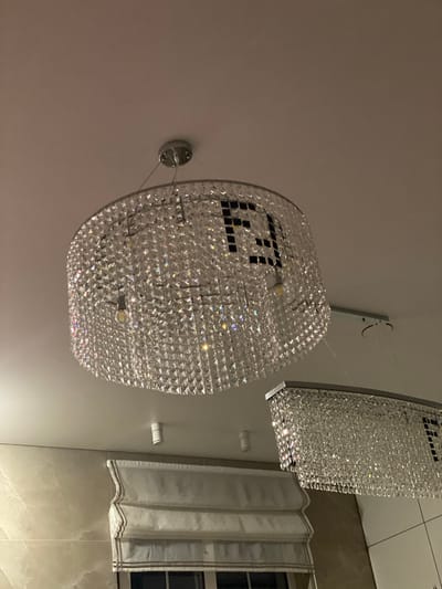 This image shows two modern crystal chandeliers hanging from a light-colored ceiling. The larger chandelier, in the foreground, is circular and appears to be made of numerous faceted crystal prisms. A dark, geometric pattern, possibly a logo or design, is visible on the inside of this chandelier. A single exposed light bulb hangs from the center of this chandelier. To the right and slightly lower, a second rectangular chandelier is visible, also adorned with crystals. Below the chandeliers, the top of a white and brown striped Roman shade window covering is visible, mounted on a wall with light beige marble-like tiles. A small white cylindrical light fixture is mounted on the ceiling to the left of the larger chandelier. The scene is dimly lit, suggesting it might be nighttime or an interior with minimal natural light. No people are present in the image. The location is specified as the city of Дорожный, Russia.