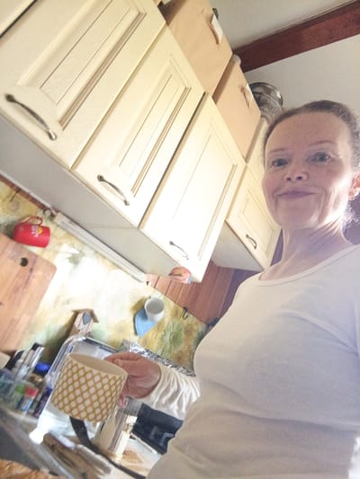 In what appears to be a domestic kitchen in Alexandrovskaya, Russia, a woman is captured in a selfie. She is wearing a plain white t-shirt and has her hair pulled back. Her expression is neutral, and she is holding a patterned yellow and white mug. The background reveals a kitchen setting with light-colored cabinets, a wooden cutting board, and various household items, including bottles and a small decorative birdhouse. A red mug is hanging on the wall. The lighting suggests it is daytime.