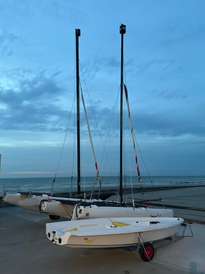 At dusk in Veules-les-Roses, France, three sailboats are positioned on a paved surface near the beach. Two identical catamarans, one behind the other, are propped up on stands. They are white with black masts and striped sails. The sails have the website "sirena-voile.com" printed on them. A smaller, single-person sailboat, also white, is in the foreground, resting on a trailer with red wheels. The sky is a deep blue with scattered clouds, and the ocean is visible in the background. The lighting suggests it is either early morning or late evening.