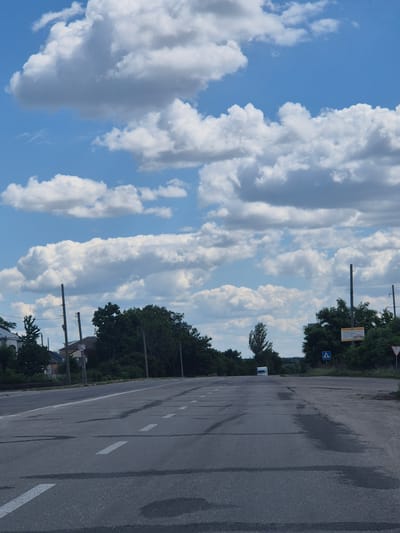 The image depicts a wide asphalt road with faded white lane markings, leading into the distance. The road surface shows signs of wear and has dark patches, possibly from recent rain or oil stains. Along the sides of the road, there are tall green trees and bushes. In the background, a white van is visible on the road. To the right, a yellow billboard is attached to a pole, and a blue pedestrian crossing sign is placed nearby. A triangular red and white sign, likely indicating a warning, is also visible to the far right. The sky above is a bright blue, dotted with numerous fluffy white clouds. The scene appears to be in a semi-rural or suburban area, consistent with the outskirts of Pidhaitsi, Ukraine. No people are present in the image.