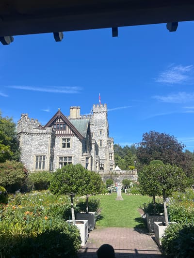 The image depicts a sunlit view of a stone castle, likely Hatley Castle, in Langford, Canada. The castle has multiple turrets and a prominent flag atop its highest tower, flying a red and white flag, possibly the Canadian flag. In the foreground, a meticulously maintained garden is visible, featuring a variety of blooming flowers in shades of orange, yellow, white, and blue, alongside manicured green shrubs and trees. A brick pathway leads towards a grassy lawn with a stone monument. The sky above is a clear, bright blue with a few wispy white clouds. A dark wooden structure, possibly part of a gazebo or pergola, frames the top of the image. The scene appears to be a tranquil, picturesque setting, typical of a historical estate or park.