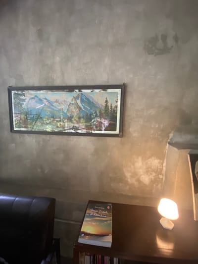 The image shows a corner of a room in a building located at -7.827, 110.482, Kecamatan Piyungan, Indonesia. The walls are concrete, with a textured, rough surface. There are several patches of lighter grey paint. On the wall is a framed artwork depicting a mountain landscape. It features a prominent mountain with snow on its peak, surrounded by pine trees and a lake in the foreground. There is a dark leather armchair to the left, and a wooden table to the right. On the table lies a book titled "Senja Indah di Kiamana". A small lamp is positioned on the table to the right of the book, casting light on the scene. The lamp has a white, round shade with a white base. The table is made of wood, with a smooth dark brown surface and a shelf underneath it. The shelf holds a stack of books that are mostly obscured from view. The room is brightly lit, suggesting it is daytime. The scene appears to be a relaxing and serene corner of a home or cafe. There are no people in the image.