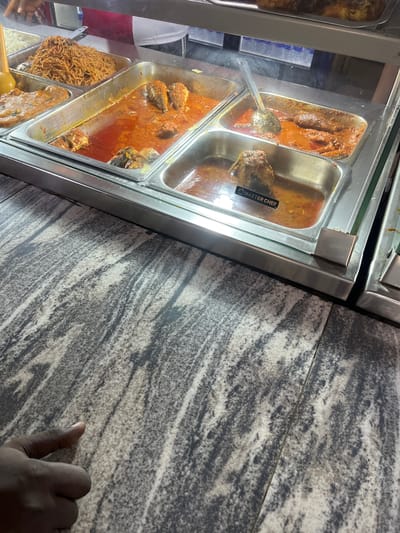 The image provides a tilted, close-up view of a food display, likely within a cafeteria or buffet setting in Osogbo, Nigeria. The scene features multiple stainless steel warming pans, commonly known as chafing dishes, filled with various dishes and covered by a clear glass shield.
In the foreground and middle, large pans hold a rich, reddish-orange stew or sauce containing sizable pieces of fish, indicating a fish stew, a common dish in Nigerian cuisine. A serving ladle is partially submerged in one of these pans. To the lower left, another pan contains a noodle dish that resembles Jollof spaghetti, characterized by its reddish-brown color and individual strands. Below it, a pan appears to hold white rice. Another pan on the far left shows a darker, perhaps meat-based, stew.
A notable detail is a black label with the text "MASTER CHEF" visible on what appears to be a food item or packaging within one of the fish stew pans.
The food display is set upon a patterned surface, possibly a granite countertop or tiled table, featuring a mix of grey, white, and dark striations. In the lower right corner, a hand with a dark skin tone is visible, resting on or near this surface, suggesting a person interacting with the food display, either waiting to be served or about to serve themselves. The lighting indicates an indoor setting, likely with artificial illumination highlighting the food. The overall scene depicts a typical ready-to-serve food counter.