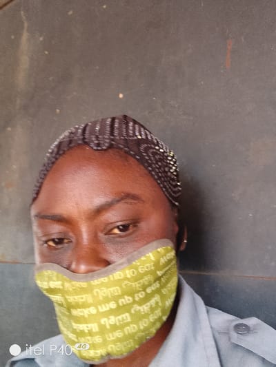 A close-up, slightly low-angle photograph depicts an individual in Hoche, Nigeria, against a plain, dark grey or dark brown textured wall. The subject has dark skin and is wearing a dark, sheer head covering adorned with reflective beads or sequins in linear and circular patterns. A light olive-green fabric face mask covers the lower half of the face, from below the eyes to the chin. The mask features repeating, inverted white or light-colored text, including the phrase "to eat," and has a lighter-colored trim. The individual's eyes are visible, looking slightly downwards, and their dark eyebrows are well-defined. A portion of a light grey or light blue-grey collared shirt with a visible buttoned shoulder tab is seen on the subject's right. A "itel P40 4G" watermark is present in the lower left corner of the image.