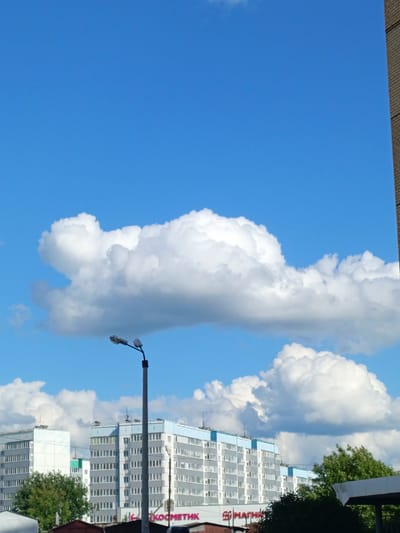The image shows a bright blue sky with large, fluffy white clouds. In the foreground, there is a street lamp with two lights. Behind the street lamp, there are several multi-story apartment buildings with light gray facades and light blue trim. Green trees are visible between the buildings. The image appears to be taken in a residential area. While no specific details in the image definitively place it in Фролы, Russia, the architectural style of the buildings is consistent with urban areas in Russia. The image captures a typical day in a city with clear weather and visible infrastructure.