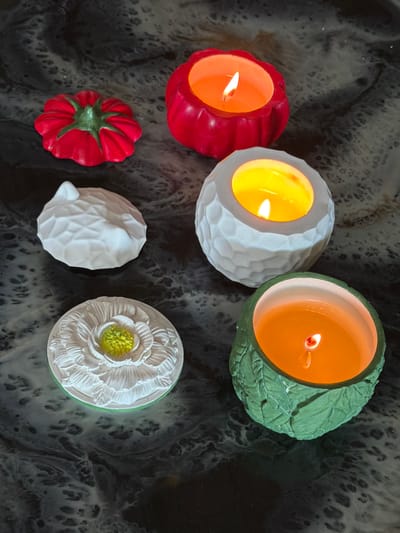 This media file showcases a collection of decorative objects, likely candles, arranged on a dark, textured surface. The subjects are primarily the candle holders and the lit candles within them. There are four lit candles in total. Two are housed in bulbous, textured holders: one red with a scalloped edge, and one white with a geometric, faceted pattern. The other two are in more ornate holders: a green one with a leaf-like texture and a white one shaped like a flower with a yellow center. In addition to the lit candles, there are two unlit decorative objects. One is a red, star-shaped item with a green stem, resembling a stylized tomato or pepper. The other is a white, egg-shaped object with a similar geometric texture to the white candle holder. The scene appears to be indoors, with the lighting suggesting artificial light or perhaps natural light from a window, though no specific time of day or weather conditions can be determined. The dark, mottled background provides a contrasting backdrop to the colorful and textured objects. There are no people or discernible text in the image. The overall impression is one of decorative arrangement, possibly for display or a cozy ambiance.