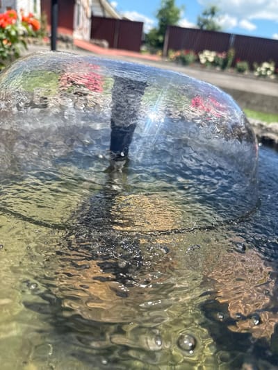 The image is a close-up, outdoor, eye-level shot of a fountain's water dome. The water is clear and rippling, with visible distortions of the background. The background is out of focus, showing a building with a reddish-brown fence and some flowers in the distance, suggesting a residential or garden setting. The sky is partly cloudy. The scene appears to be a sunny day. No people are visible. The location is Hraniv, Ukraine.