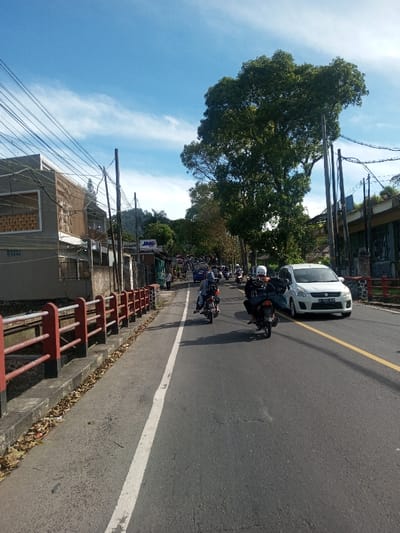 The image displays a sunlit street scene, likely in an urban or semi-urban area, observed from a low angle. The asphalt road features a solid white line on its left edge and a double yellow line further right, indicating lane demarcation. Traffic consists primarily of motorcycles and at least one white multi-purpose vehicle (a Suzuki Ertiga with Indonesian license plate B 1255 SJQ).

In the immediate foreground, two motorcycles are visible, traveling away from the camera. The motorcycle on the left carries two riders; the passenger is wearing a light blue head covering. The motorcycle on the right has a single rider wearing a white helmet and a dark jacket, with a dark item secured behind them. A line of other vehicles, predominantly motorcycles, extends into the distance, indicating ongoing traffic.

To the left of the road, a narrow concrete strip, possibly a sidewalk, is lined by a low barrier with red horizontal rails and black posts. Dried leaves are scattered on this strip. Behind the barrier, a multi-story building with grey walls and a decorative patterned element on its upper floor is visible. Further down the street, a building displays a "JNE" sign, a courier company originating from Indonesia.

The right side of the street is lined with utility poles supporting numerous overhead power lines. A large, mature tree with dense green foliage stands prominently in the midground on the right, with additional trees extending along the street. Distant hills or mountains are visible in the background. The sky is clear blue with sparse, wispy white clouds, and direct sunlight illuminates the scene, casting shadows on the roadway.