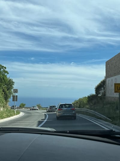 The image captures a view from inside a vehicle driving on a road in Vrela, Montenegro. The road curves to the left, with a steep, vegetated embankment on the left and a stone-faced building with a large "FOR" sign on the right. Several cars are visible on the road ahead. The sky is a bright blue with wispy white clouds, and the sea is visible in the distance. The overall scene suggests a sunny day with moderate traffic on a scenic coastal road.