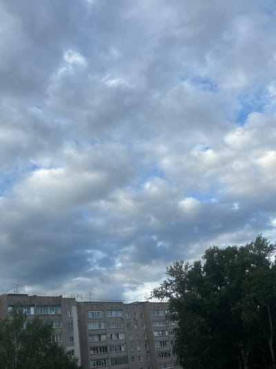 This media file features an upward-looking view of a cloudy sky with glimpses of blue, partially obscured by a tall, grey, multi-story building on the right side. A portion of a green tree is visible in the upper right and lower right corners. The building has numerous windows, some of which appear to be open. Scaffolding is also visible on the side of the building. The overall impression is of an urban setting under a potentially overcast sky, suggesting it could be daytime. The image does not contain any visible text or people, nor does it depict a specific activity or event. The location is identified as Kirov, Russia.