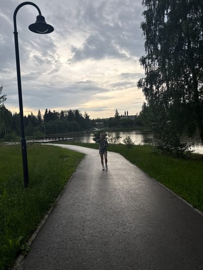 In Chusovoy, Russia, an individual is captured in a full-body shot, walking on a paved path that curves towards the viewer. The person, dressed in shorts and a hooded sweatshirt, faces the camera, their hands near their face, appearing to be speaking or gesturing. They are mid-stride on the wet asphalt path, which is bordered by lush green grass on the left and a large body of water on the right.

The water reflects the overcast sky, characterized by dramatic clouds with streaks of sunlight breaking through. A dark lamp post stands prominently on the left side of the frame, silhouetted against the sky. In the background, a dense line of trees forms a backdrop to the lake, and beyond the trees, in the distance, two distinct towers of a building can be seen. The overall scene suggests an outdoor setting in a park or recreational area during what appears to be late afternoon or early evening.