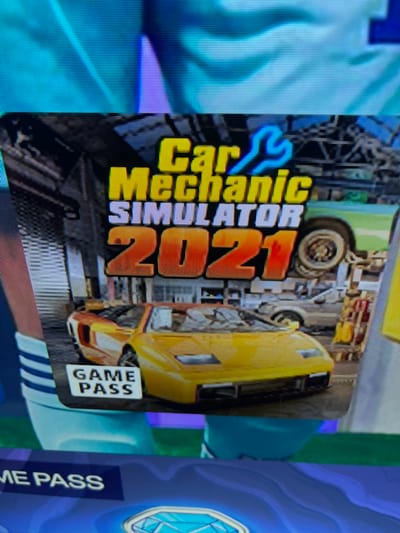 The media file displays a screen showing a video game cover. The cover features a yellow sports car, possibly a Lamborghini, partially disassembled, with its engine exposed. The background appears to be a garage or workshop setting with tools and car parts. The text "Car Mechanic Simulator 2021" is prominently displayed in a vibrant yellow and orange font. Below this, a white rectangle with the words "GAME PASS" in blue lettering is visible, indicating the game is available on the Game Pass service. To the right of the game cover, other game-related graphics are partially visible, including a blue background with a wave-like pattern and a circular emblem. The lighting suggests an indoor setting, likely a digital display. There are no people visible, and the focus is on the game's artwork and branding. The overall impression is that of a promotional image for a racing or simulation video game.