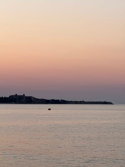 The image depicts a serene coastal scene at what appears to be sunset or sunrise, with a gradient of soft pink and peach hues filling the sky. A calm sea occupies the lower half of the frame, reflecting the muted colors of the sky. In the distance, a peninsula or a long stretch of coastline is visible, featuring a cluster of buildings that form a small town or village. To the left of the town, a large, multi-story building with a distinct architectural style is prominent. Further along the coastline, a small lighthouse or tower-like structure can be observed. A single, bright red buoy floats on the water, positioned approximately in the center of the frame. The overall atmosphere is tranquil and still, suggesting a peaceful moment. The location is identified as Ravda, Bulgaria.