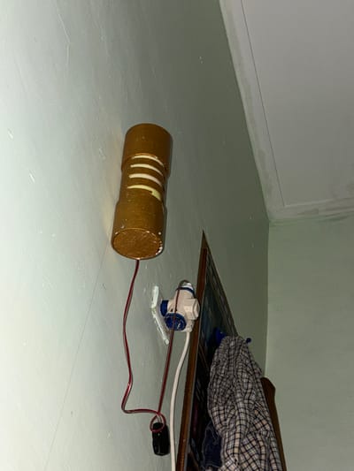 A homemade, cylindrical lamp with horizontal stripes of light and dark gold is mounted on a light green wall. A red wire extends from the bottom of the lamp to a small black object, possibly a transformer. Below the lamp, a white electrical socket is mounted on the wall, with a blue and white connector plugged into it. Two wires run from the socket, one red and one white. To the right of the electrical socket, a framed object, possibly a mirror or a picture, leans against the wall. A plaid shirt is draped over the top of the framed object. The ceiling is visible in the upper right corner of the image and appears to have water stains. The image is taken from a low angle, looking upwards towards the lamp and ceiling. The location is identified as Tupah, Indonesia.
