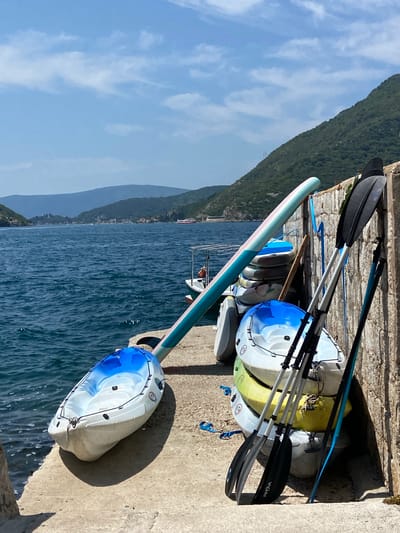 In Perast, Montenegro, on a sunny day, a concrete pier is laden with various watercraft and paddles. A long, pale blue and pink paddleboard rests diagonally across the pier, its tip near a white and blue kayak positioned at the water's edge. Behind these, a stack of four kayaks, in white, yellow, and light blue hues, leans against a rough stone wall. Three paddles, two with black blades and silver shafts and one with a blue shaft and black blade, stand upright beside the stacked kayaks. The deep blue waters of the Bay of Kotor stretch out to the horizon, with verdant, tree-covered hills rising in the background. A small boat with a white canopy is visible to the left, and distant buildings and mountains dot the landscape. The scene is devoid of people.
