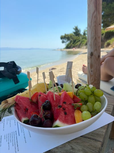 This is a close-up, rotated image of a fruit platter served in a white bowl, set on a rustic wooden table. The platter contains an assortment of fresh fruits, including green grapes, watermelon slices, pineapple chunks, cherries, and strawberries, all skewered with small wooden picks. In the background, a beach scene unfolds: a sandy shore meets the calm, turquoise water of the sea under a clear blue sky, suggesting a bright, sunny day, likely late morning or early afternoon. A person's legs and feet are partially visible in the upper background, indicating they are seated near the table, perhaps enjoying a meal or relaxing. The setting appears to be a beachside cafe or restaurant in Kassandra Municipal Unit, Greece, given the menu visible in the foreground. The menu lists various food and drink options, including "Agora Beach | Premium Family Combo" and "Kid's Beach Snack," confirming the location's name and its likely purpose as a leisure establishment. The overall mood is relaxed and summery.