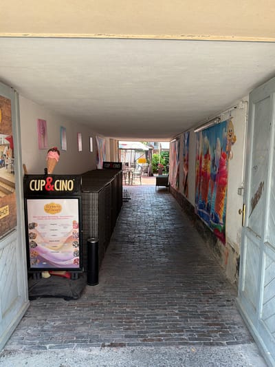 The image depicts a narrow alleyway with brick paving, likely in the city of Wismar, Germany. On the left side, a sign for "Werners Eiscafe" is displayed, featuring colorful images of ice cream and menu items. Adjacent to the sign is an outdoor display of a large, decorative ice cream cone. Further down the alley, large, vibrant posters with images of ice cream are hung on the wall. The alley leads to an outdoor seating area with tables and chairs, suggesting a cafe or restaurant. The overall scene is that of a commercial establishment in a European city.