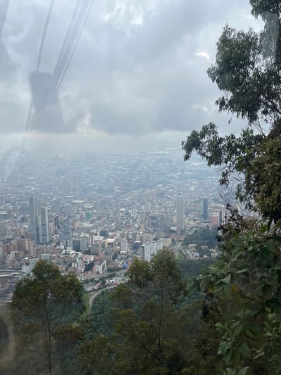 This image provides an elevated, panoramic view of the expansive city of Bogotá, Capital District, Colombia, likely captured from the Monserrate Funicular or Teleferico system. The scene is observed through a glass surface, evidenced by distinct reflections of vertical cables and support structures in the upper left quadrant.

The cityscape dominates the midground and background, extending outwards under a heavily overcast and hazy sky. Numerous buildings of varying heights populate the urban sprawl; prominent high-rise structures are visible in the mid-left area, characteristic of Bogotá's financial district. The city's density is apparent, with residential and commercial buildings tightly packed. Roads and urban infrastructure are discernible, though details are obscured by atmospheric haze.

In the foreground, the immediate surroundings consist of dense, verdant foliage, including trees with dark green leaves and various shrubs, indicating a mountainous or steeply sloped terrain. No individuals are depicted, and no specific actions or interactions are occurring within the visible frame. The overall environment is one of an urban metropolis set against a natural, elevated landscape, viewed under diffuse light conditions due to the cloudy sky.