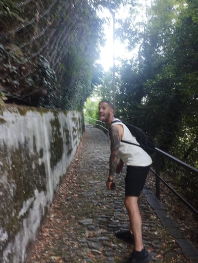 In Orselina, Switzerland, a man with tattoos on his arms and a white tank top is walking up a cobblestone path lined by a concrete wall on the left and trees on the right. He has black shorts and is wearing a black backpack. His tongue is sticking out, and he is looking back over his left shoulder. The path is covered with fallen leaves, and there is a metal railing on the right side of the path. The sun is shining through the trees, creating a bright, backlit effect.