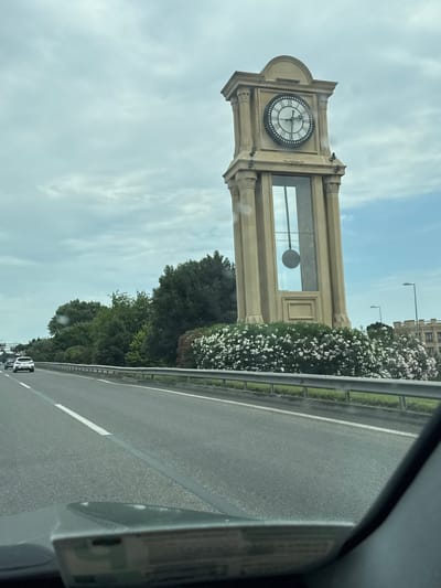 The image displays a large clock tower beside a highway. The clock face is visible, showing Roman numerals and hands indicating a time around 3:15. The clock tower itself is made of light-colored stone and has architectural details like columns and decorative moldings. Adjacent to the tower, a dense green bush with white flowers is growing, bordering the highway.

The scene appears to be taking place during the daytime, with a bright sky and visible clouds, suggesting overcast or partly cloudy weather. The highway is paved and has white lane markings. A metal guardrail separates the road from the landscaped area. In the distance, several streetlights are visible, and a building with multiple windows can be seen above the trees and bushes.

The perspective suggests the photo was taken from a moving vehicle on the highway, looking towards the clock tower. The angle is slightly tilted, indicating the vehicle is not perfectly aligned with the road. No people are clearly visible in the frame, but the presence of the highway implies ongoing traffic. The overall impression is a roadside view of a prominent architectural landmark. The location is identified as Surakhany Raion, Azerbaijan.