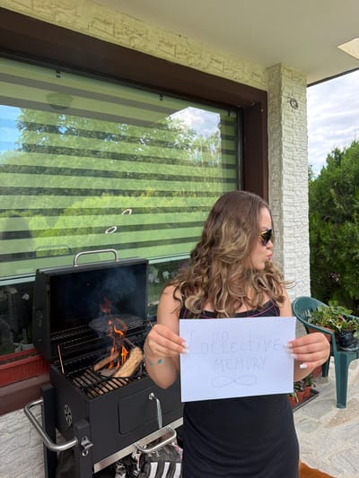 In Priseltsi, Bulgaria, a woman stands outdoors in front of a grill with a fire going. She is holding a piece of paper with the words "collective memory" and an infinity symbol drawn on it. The woman is wearing a black dress and sunglasses. The background shows a house with a stone facade and a window with blinds. There are also trees and plants visible. The overall setting appears to be a backyard or patio area.