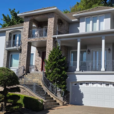 This is a wide, eye-level, exterior shot of a multi-story, modern house. The house is predominantly grey with stone accents. The main facade is stone with a central entrance and a two-story balcony above it, supported by stone pillars. There are multiple windows throughout the house, some with balconies and ornate railings. A large, grey garage door is visible on the right side of the house. The landscaping includes a manicured, topiary-like tree on the left, and trimmed hedges. A clear blue sky is visible above the house. The image appears to be taken during the daytime. The location is a residential area in Portland, United States of America.