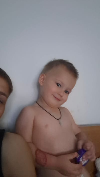The media file shows a close-up, rotated shot of a baby and a portion of an adult's torso. The baby is the primary subject, with their torso and head visible. They appear to be looking towards the viewer with a slight smile. The baby is wearing a thin, dark necklace. On the baby's left arm, there is a red, textured mark or dressing. The baby's hands are holding a small, purple object with some white writing on it. The adult's torso, wearing a dark garment, is visible in the upper left of the frame, suggesting the baby is being held. The background is a plain, light-colored wall. The overall lighting suggests it might be daytime, possibly indoors. There is no discernible weather information. The location context is Zielona Góra, Poland.
