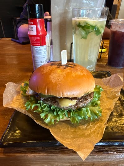 An overhead shot captures a close-up of a gourmet burger served on crumpled brown paper atop a dark, triangular plate. The burger features a golden-brown bun, a thick beef patty, melted cheese, and a generous bed of crisp green lettuce. A small wooden skewer pierces the top bun. In the background, out of focus, are two drinks: a tall glass filled with a pale yellow liquid and ice, with cucumber slices and mint leaves visible, and a smaller cup containing a dark red beverage. To the left, a red squeeze bottle with a black cap is partially visible, with white text and a black barcode on its label. The setting appears to be a restaurant table, likely in Manzherok, Russia, given the context of the prompt, with a wooden surface beneath the plate.