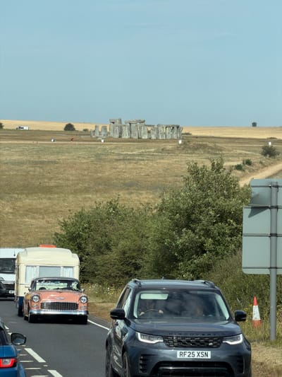 The image shows a road with several vehicles traveling on it. In the foreground, a dark gray SUV is visible, with the license plate "RF25 XSN." To the left of the SUV, a portion of a blue car is visible. Further down the road, a vintage orange car is pulling a caravan. The caravan has a beige exterior with a white roof and a dark blue stripe along the bottom. Behind the orange car, a white van is also on the road. In the background, the Stonehenge monument is visible in a field. The sky is clear and blue. The scene appears to be in a rural area, likely in or near West Amesbury, United Kingdom, given the presence of Stonehenge.