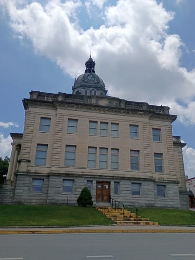 The image shows the exterior of a historical courthouse building with a prominent clock tower. The courthouse is constructed of stone and has multiple windows. A set of yellow stairs with black railings leads up to the main entrance. The building is situated on a grassy slope, with a road in the foreground. The sky is partly cloudy. Based on external knowledge, this is likely the Henry County Courthouse located in Paris, Tennessee, United States of America, as it is a well-known landmark in that city.