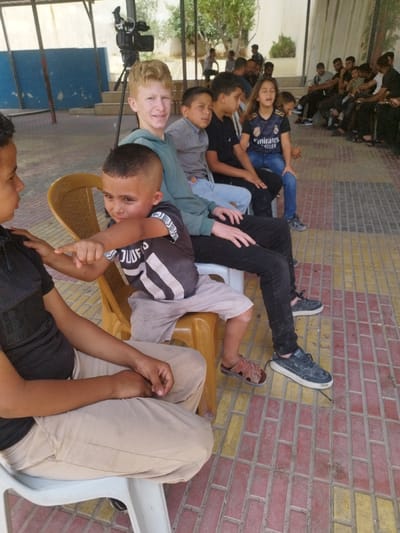 This image depicts an outdoor gathering in Idhna, Palestinian Territory, under a light-colored shaded structure supported by metal poles. The ground is paved with rectangular bricks in alternating red-brown and yellow patterns.

In the foreground, six individuals, primarily children and adolescents, are seated in a row on plastic chairs. From left to right:
1.  A male, visible from the torso down, wearing a dark shirt and light-colored pants, seated on a white chair.
2.  A young boy with a short haircut, wearing a dark striped t-shirt and light shorts, is seated on a yellow chair, his left foot clad in a brown sandal. He is leaning slightly right, with his right hand on the shoulder of the adjacent individual, and his left arm extended, pointing his index finger to the left.
3.  A fair-haired male adolescent in a green hooded top and black pants, seated on a white chair. He is looking directly at the camera and smiling. The young boy from position 2 is partially on his lap.
4.  A boy with dark hair, wearing a light gray long-sleeved shirt and light blue jeans, seated on a white chair, looking towards the right.
5.  A boy with dark hair, wearing a black t-shirt and dark pants, seated on a white chair, looking towards the right.
6.  A girl with long dark hair, wearing a dark blue Real Madrid jersey and blue jeans, seated on a white chair, looking towards the right with her eyes closed or cast downwards.

In the mid-ground, slightly to the left and behind the seated group, a black video camera is mounted on a tripod. In the background to the right, approximately 10-15 additional individuals, mainly males of various ages, are seated or standing, appearing to observe something out of frame. A blue wall is visible in the distant background to the left, with some green foliage and structures beyond the immediate shaded area. The scene is illuminated by natural daylight.