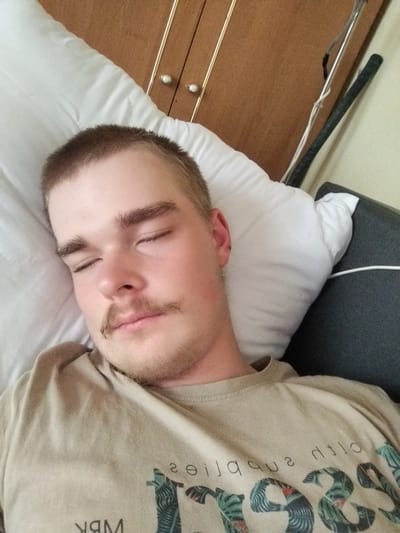 A young person with short brown hair and a light mustache is sleeping on a white pillow, with their head turned to the side. They are wearing a tan t-shirt with a graphic design on it. The background shows a wooden cabinet and a portion of a dark gray surface, possibly a couch or chair. The scene appears to be indoors, in a residential setting. There is no identifiable information linking this image to Cēsis, Latvia.