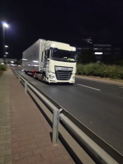 A white DAF semi-trailer truck travels at night on a multi-lane road. The truck is moving from left to right in the frame, and the image is blurred due to motion. The truck has a white cab with a silver trailer. It is illuminated by streetlights, with one prominent streetlight on the left side of the road. The road is paved and has a white lane marking. To the left of the road is a pedestrian walkway made of brick pavers, separated from the road by a metal guardrail. Behind the guardrail and walkway, there is a line of green bushes and trees. In the background, several multi-story buildings with illuminated windows are visible, suggesting an urban or suburban setting within Frankfurt, Germany. The sky is dark, indicating it is nighttime.