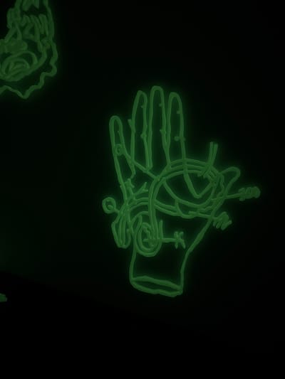 A luminescent, crude drawing of a hand dominates the upper portion of the image against a black background. The hand appears to be holding or interacting with an assortment of chaotic, squiggly lines and geometric shapes. In the lower left corner, there's a partially visible clump of similar luminescent lines. The overall aesthetic evokes a sense of amateur artistry in a neon or glow-in-the-dark medium.