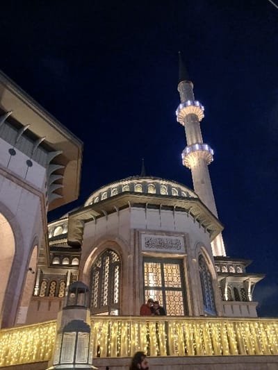 A nighttime view of the exterior of the Eyüp Sultan Mosque in Istanbul, Türkiye (latitude: 41.037, longitude: 28.984). Two men stand on a balcony facing the viewer, and the balcony is decorated with white string lights. The mosque is lit up, the minaret is tall and narrow, and the sky is dark. The mosque is lit up, and the minaret is tall and narrow. The sky is dark, and the only lights visible are the streetlights. There is a large window in the center of the mosque, and intricate detailing on the building. The mosque is located in the Eyüp district of Istanbul. It is a popular destination for Muslim pilgrims and tourists alike.